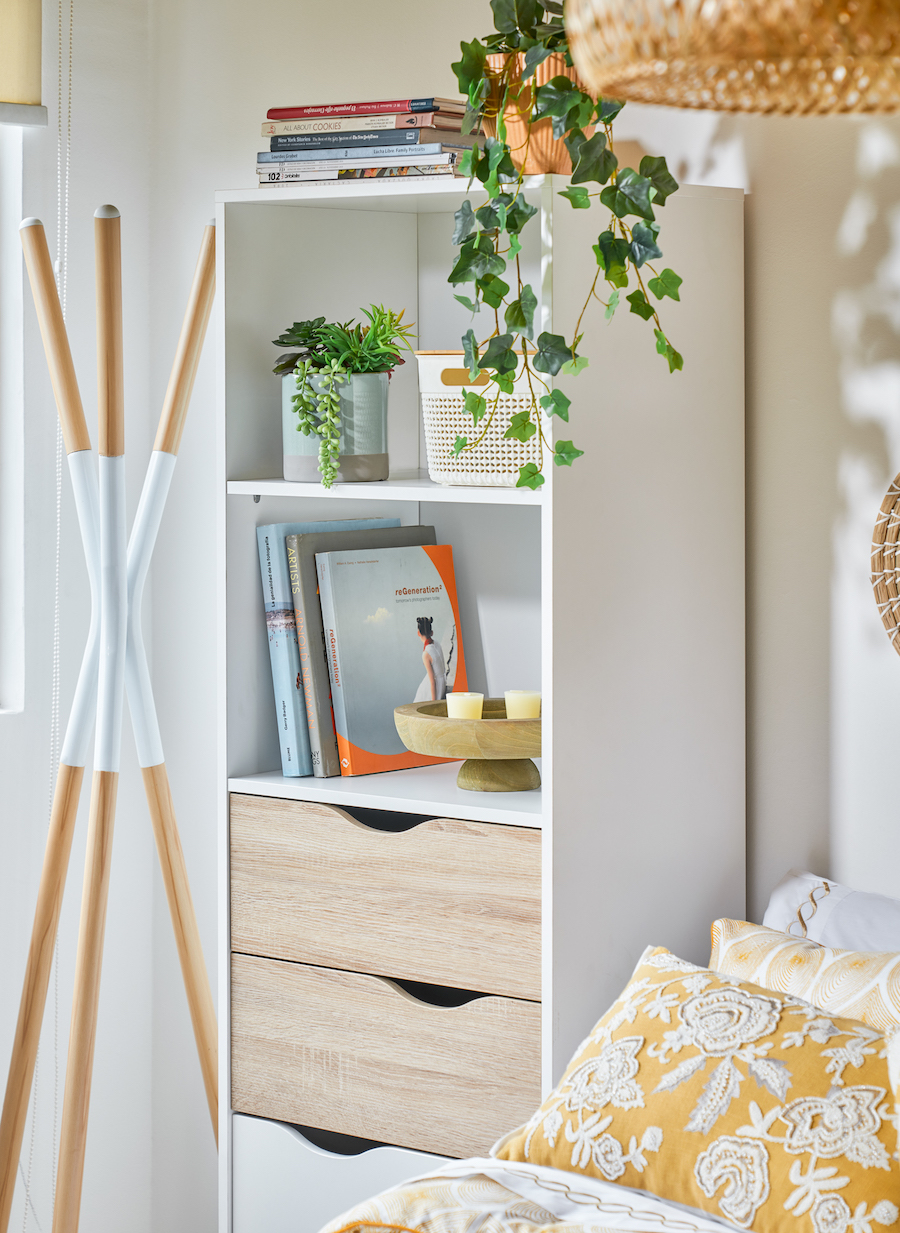 Dormitorio con estantería blanca con repisas con libros y plantas y cajones inferiores. A su lado hay una cama y al otro lado un colgador de ropa de pie.