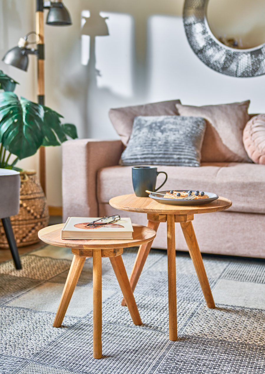 Living estilo nórdico con sofá rosa y cojines grises. Set de mesas de apoyo de madera, redondas. Gran planta en canasto y pouf gris