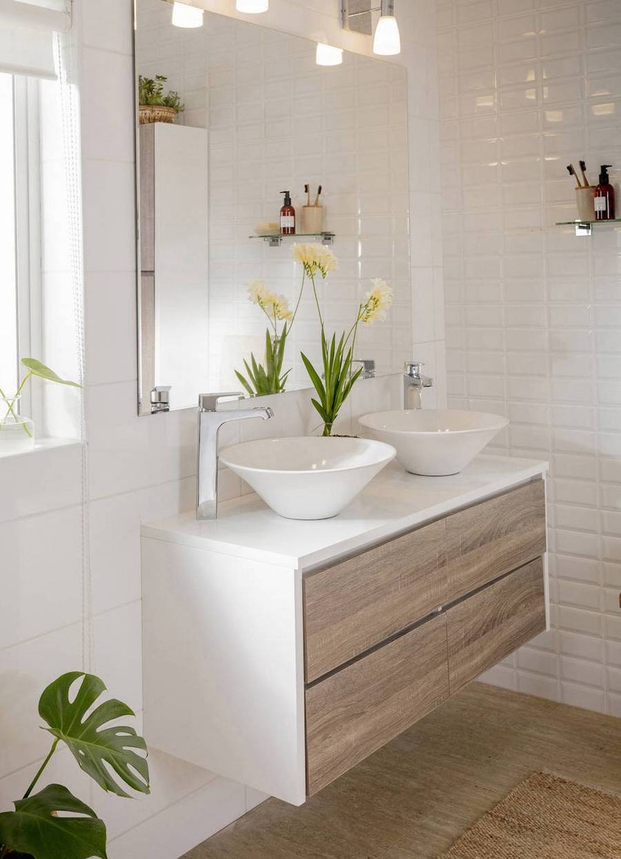 Baño de cerámicas blancas con un mueble vanitorio flotante blanco, con cajones de madera y 2 lavamanos separados del mismo mueble. La grifería es plateada y hay unas flores amarillas que decoran el espacio.
